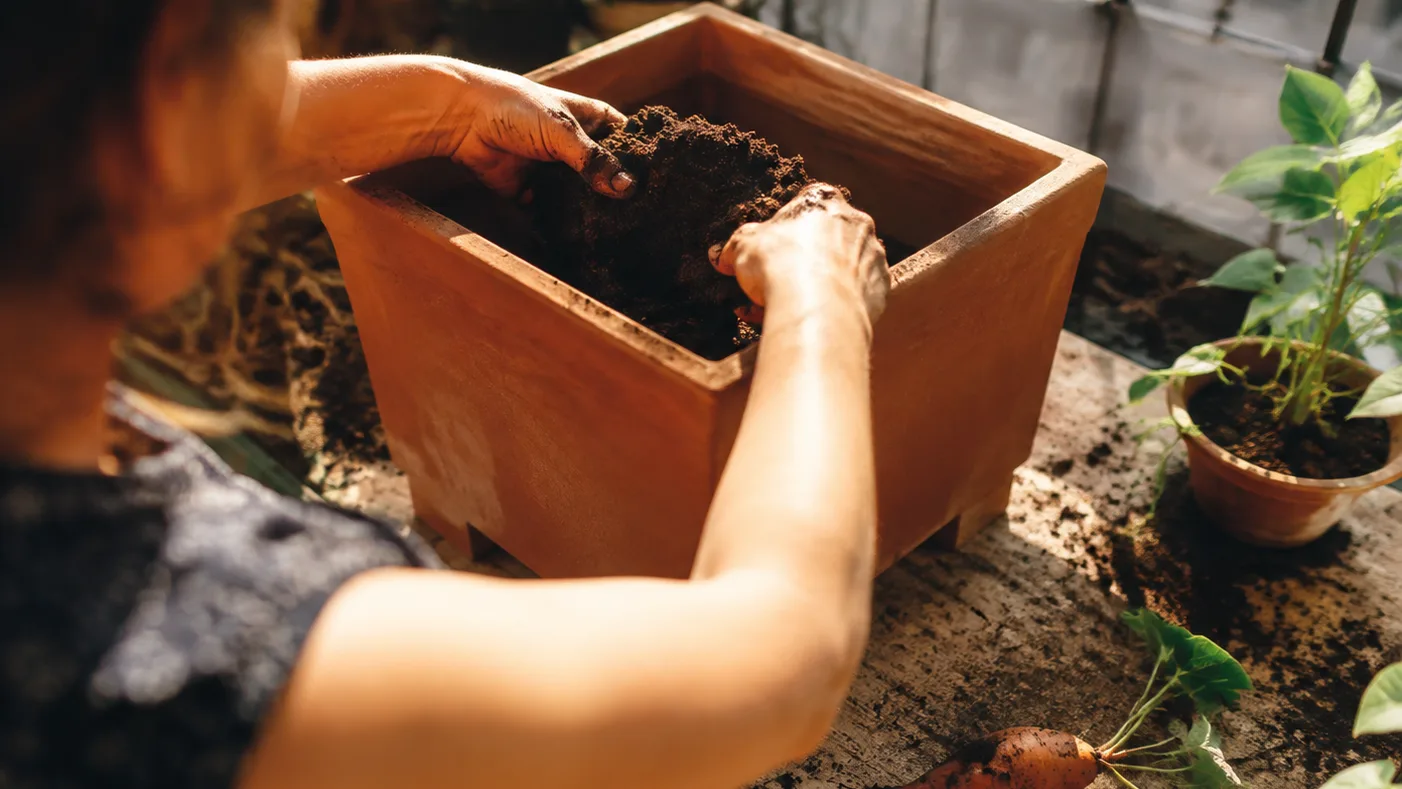 Hands fill a raised square planter with loose mix while its drainage holes stay open beneath it.
