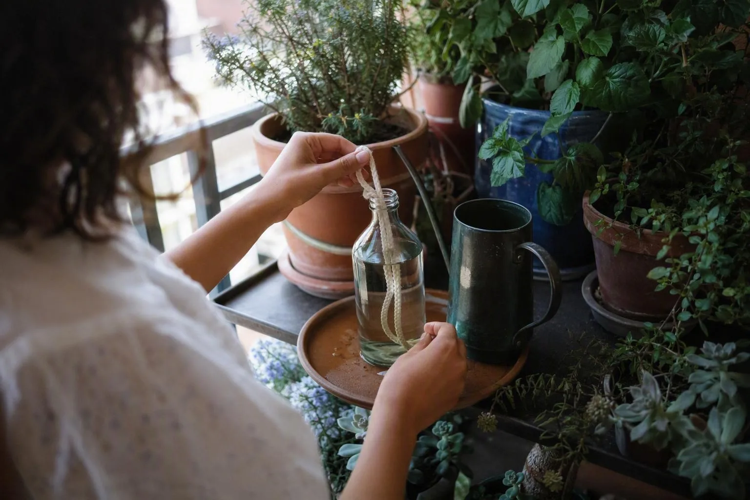 Hands arrange simple watering supplies beside potted balcony plants in bright shade for a trip away.